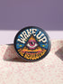 A round pinback button featuring a retro psychedelic design. The central focus is a stylized Eye of Providence in a pink triangle, surrounded by vibrant orange and yellow flames. Behind the triangle are teal and petrol blue sunburst rays. The text "WAKE UP" curves across the top in cream wavy letters, and "THE REVOLUTION" curves along the bottom in orange. The button has a glossy finish and is available in 1.75 inch or 2.25 inch sizes.