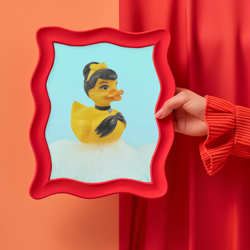 A chic rubber duck styled like Audrey Hepburn’s Holly Golightly, wearing a high bun, yellow bow, black gloves, and a sparkly choker. The duck floats in a pile of foam and is framed in white on a bubbly bathroom shelf with a red tile backdrop and soap dispenser.