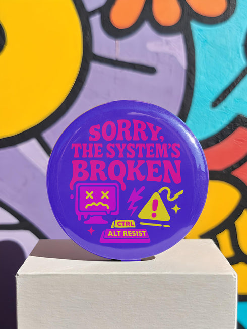 A vibrant purple pinback button with a glossy finish. The main text reads "SORRY, THE SYSTEM'S BROKEN" in a chunky, dripping pink font. Below the text are several retro tech icons in pink and yellow. On the left is a glitching 90s-style computer monitor with a sad face and X's for eyes. In the center is a yellow warning sign with an exclamation point, a lightning bolt, and sparkles. Below is a keyboard command parody that reads "CTRL ALT RESIST.