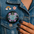 Glossy black pinback button featuring embroidered-style artwork of a pastel purple heart that reads “Read or Die” pierced by a sword and surrounded by roses; shown on books, graffiti, and lace backgrounds.