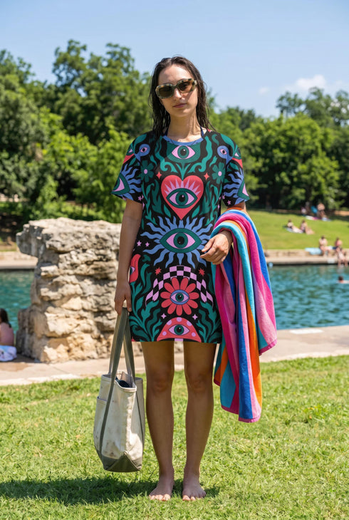 A short-sleeve, above-the-knee t-shirt dress with a true black background. The front features a vibrant, vertically stacked symmetrical graphic design. From top to bottom, the design includes a pink heart containing a green and blue eye, a large blue sunburst with a central eye, a red daisy flower with an eye flanked by pink and black checkered arches, and a pink mushroom cap with an eye. Swirling emerald green foliage, pink stars, and purple stars fill the negative space around the motifs.