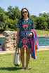 A short-sleeve, above-the-knee t-shirt dress with a true black background. The front features a vibrant, vertically stacked symmetrical graphic design. From top to bottom, the design includes a pink heart containing a green and blue eye, a large blue sunburst with a central eye, a red daisy flower with an eye flanked by pink and black checkered arches, and a pink mushroom cap with an eye. Swirling emerald green foliage, pink stars, and purple stars fill the negative space around the motifs.