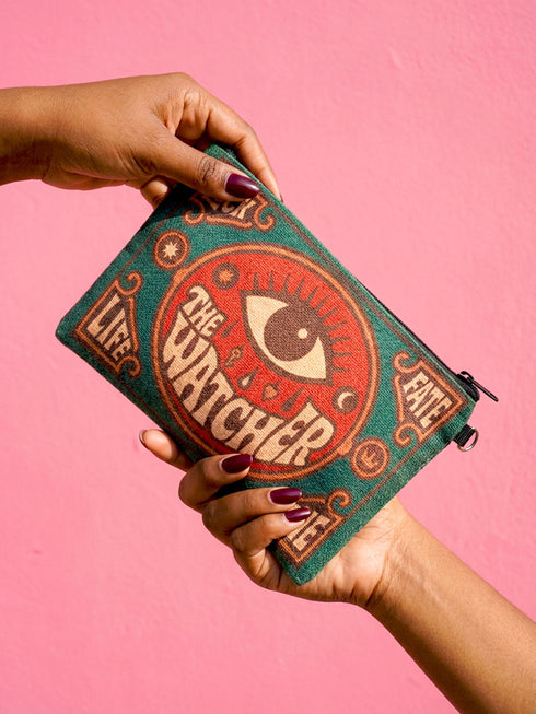 A rectangular 9x6 inch makeup bag and zipper pouch featuring a textured linen-like polyester fabric. The bag showcases a bold, maximalist print with vivid colors typical of retro 70s and pastel goth aesthetics, centering on a large mystic eye design labeled "The Watcher." It features a sturdy black zipper across the top and includes a removable black nylon wristlet strap.Perfect for use as a pencil case, cosmetic organizer, or festival wristlet.
