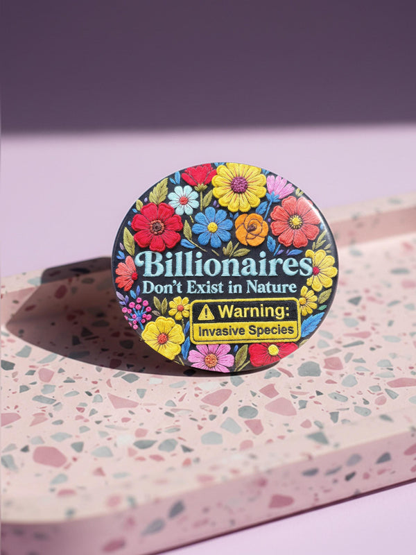 A round pinback button featuring a flat-printed design that mimics the appearance of highly detailed, 3D embroidery (trompe-l'œil). The background is solid black, with a floral border including red, yellow, sky blue, magenta, and orange flowers. In the center, the text "Billionaires Don't Exist in Nature" is written in a blue serif font. Below the text is a yellow rectangle with black borders containing a warning icon and the words "Warning: Invasive Species" in a bold, sans-serif black font.