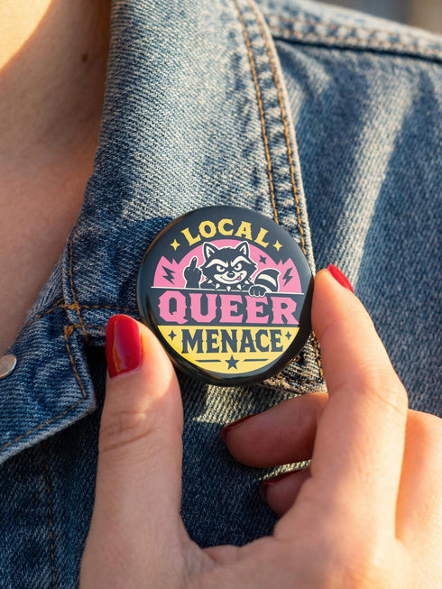 Round pinback button badge with Local Queer Menace text in bold yellow and pink retro lettering on a black background. A cartoon raccoon with a spiked collar flips the middle finger with a painted pink nail, framed by a hot pink sunburst, lightning bolts, and star accents. Glossy laminate finish over durable metal tinplate with a secure safety pin backing. Funny queer pride pin available in 2.25 inch and 3 inch sizes for jackets, totes, lanyards, and backpacks.