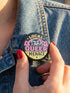 Round pinback button badge with Local Queer Menace text in bold yellow and pink retro lettering on a black background. A cartoon raccoon with a spiked collar flips the middle finger with a painted pink nail, framed by a hot pink sunburst, lightning bolts, and star accents. Glossy laminate finish over durable metal tinplate with a secure safety pin backing. Funny queer pride pin available in 2.25 inch and 3 inch sizes for jackets, totes, lanyards, and backpacks.