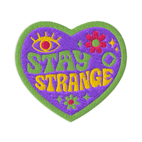 "A detailed embroidered patch in the shape of a purple heart with a vibrant green border. In a groovy, psychedelic font, the words "STAY STRANGE" are stitched in yellow and green. The design includes a mystical third eye at the top, and small retro flowers and stars, giving it a 70s hippie vibe.