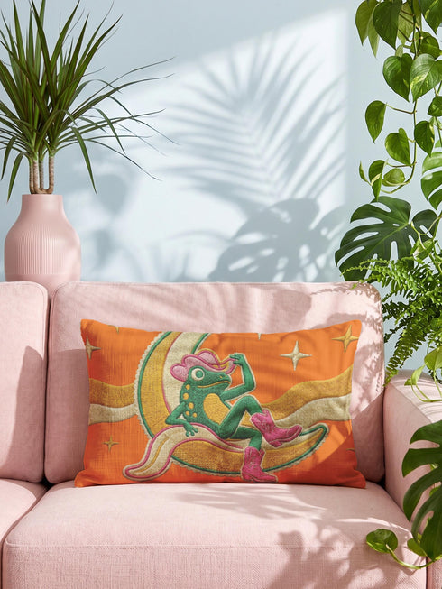 Rectangular 20x12 inch lumbar pillow cover in bright orange featuring printed faux embroidery of a green frog wearing a pink cowgirl hat and pink cowboy boots, lounging on a golden yellow crescent moon shaped like a peeled banana, surrounded by cream and gold sparkle stars and retro wavy stripes. Whimsical cowboy frog design printed on both sides with a hidden zipper, crafted from soft linen look polyester in a funky 70s retro style.