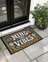 A rectangular floor mat with a dark charcoal black background. The center features the phrase 'MIND THE VIBES' in stylized, wavy cream and golden-mustard retro typography. The text is surrounded by symmetrical motifs: a golden evil eye with sun rays on the top left, a golden crescent moon with a four-point star on the top right, and two cream daisy flowers with golden stems at the bottom corners. The bottom edge has a wavy golden checkerboard pattern. A faux-stitch border frames the art.