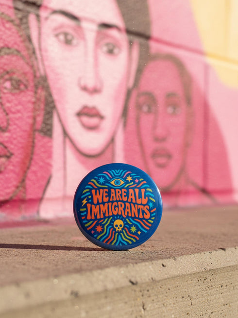 A round 1.75 inch blue pinback button featuring retro psychedelic artwork. The text reads We Are All Immigrants in a groovy orange and red 1970s font style centered on the badge. A mystic third eye sits above the text while a small skull sits below it. Wavy rainbow lines in cyan, green, and orange radiate outward from the center along with small starbursts and lightning bolts. The button has a glossy finish reflecting light.