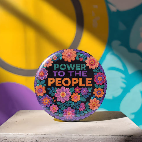 Embroidered-style pinback button featuring the phrase "POWER TO THE PEOPLE" in bold, multicolored satin-stitch letters—green, purple, and orange—framed by a vivid circular floral wreath. Pink, red, and purple flowers with leafy greens radiate around the text. Design has a handcrafted, folk-art feel with a transparent background.