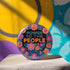 Embroidered-style pinback button featuring the phrase "POWER TO THE PEOPLE" in bold, multicolored satin-stitch letters—green, purple, and orange—framed by a vivid circular floral wreath. Pink, red, and purple flowers with leafy greens radiate around the text. Design has a handcrafted, folk-art feel with a transparent background.