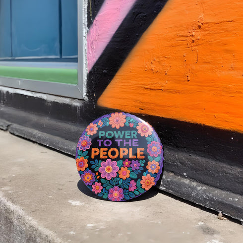 Embroidered-style pinback button featuring the phrase "POWER TO THE PEOPLE" in bold, multicolored satin-stitch letters—green, purple, and orange—framed by a vivid circular floral wreath. Pink, red, and purple flowers with leafy greens radiate around the text. Design has a handcrafted, folk-art feel with a transparent background.