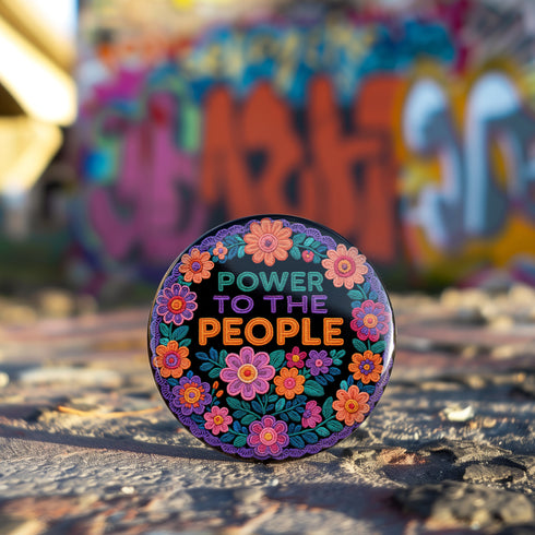 Embroidered-style pinback button featuring the phrase "POWER TO THE PEOPLE" in bold, multicolored satin-stitch letters—green, purple, and orange—framed by a vivid circular floral wreath. Pink, red, and purple flowers with leafy greens radiate around the text. Design has a handcrafted, folk-art feel with a transparent background.