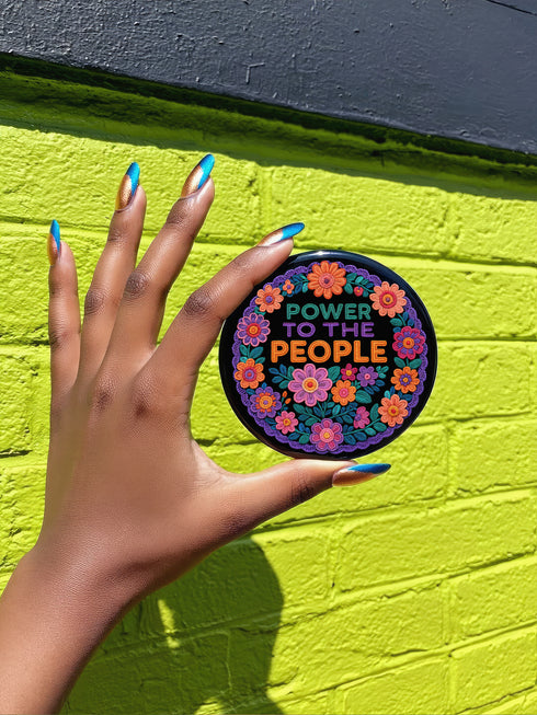Embroidered-style pinback button featuring the phrase "POWER TO THE PEOPLE" in bold, multicolored satin-stitch letters—green, purple, and orange—framed by a vivid circular floral wreath. Pink, red, and purple flowers with leafy greens radiate around the text. Design has a handcrafted, folk-art feel with a transparent background.