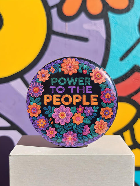Embroidered-style pinback button featuring the phrase "POWER TO THE PEOPLE" in bold, multicolored satin-stitch letters—green, purple, and orange—framed by a vivid circular floral wreath. Pink, red, and purple flowers with leafy greens radiate around the text. Design has a handcrafted, folk-art feel with a transparent background.