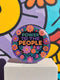 Embroidered-style pinback button featuring the phrase "POWER TO THE PEOPLE" in bold, multicolored satin-stitch letters—green, purple, and orange—framed by a vivid circular floral wreath. Pink, red, and purple flowers with leafy greens radiate around the text. Design has a handcrafted, folk-art feel with a transparent background.