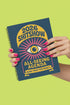 A hardcover spiral planner for 2026 with a dark blue cover and retro 70s style graphics. The title reads "2026 SHITSHOW All-Seeing Agenda" in bold yellow and pink text. Below the title is a large, stylized all-seeing eye with pink rays emanating from it. At the bottom, a pink ribbon contains the text "BLINK TWICE FOR MERCY." The back cover is also dark blue with a yellow sun graphic and the text "Plans bloom. Deadlines hum. I write, I wander, then get it done. Keep receipts. Keep grace.