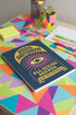 A hardcover spiral planner for 2026 with a dark blue cover and retro 70s style graphics. The title reads "2026 SHITSHOW All-Seeing Agenda" in bold yellow and pink text. Below the title is a large, stylized all-seeing eye with pink rays emanating from it. At the bottom, a pink ribbon contains the text "BLINK TWICE FOR MERCY." The back cover is also dark blue with a yellow sun graphic and the text "Plans bloom. Deadlines hum. I write, I wander, then get it done. Keep receipts. Keep grace.