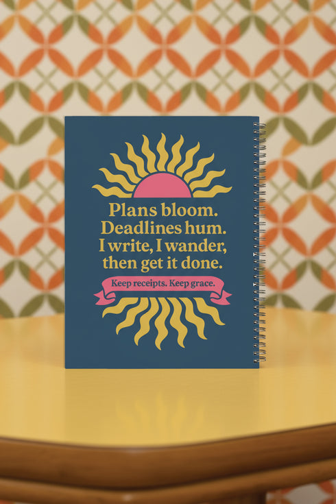 A hardcover spiral planner for 2026 with a dark blue cover and retro 70s style graphics. The title reads "2026 SHITSHOW All-Seeing Agenda" in bold yellow and pink text. Below the title is a large, stylized all-seeing eye with pink rays emanating from it. At the bottom, a pink ribbon contains the text "BLINK TWICE FOR MERCY." The back cover is also dark blue with a yellow sun graphic and the text "Plans bloom. Deadlines hum. I write, I wander, then get it done. Keep receipts. Keep grace.