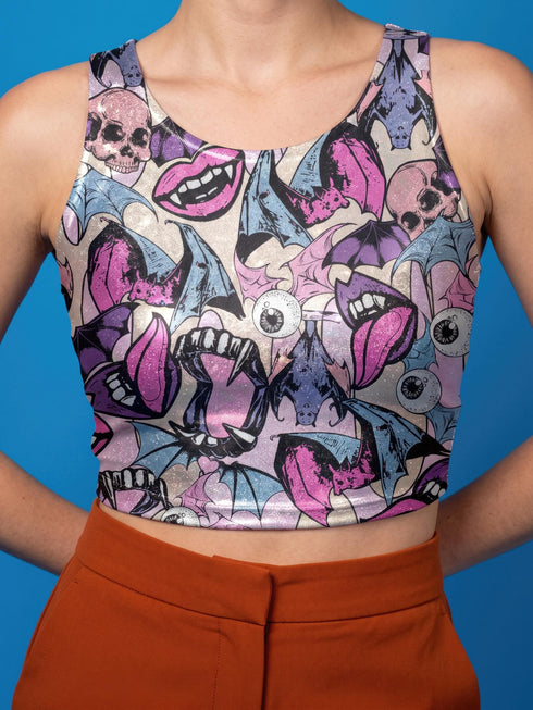 A sleeveless cropped tank top made from stretchy performance knit fabric. The all-over print features a dense, scattered pattern of pastel goth and creepy cute motifs over a light background. Graphics include pink and blue bats in flight, floating eyeballs with blue irises, pink skulls, and glossy vampire lips with sharp white fangs and tongues sticking out. The neckline is a high scoop and the armholes are curved. The fabric comes in either a matte finish or a sparkly silver glitter foil.