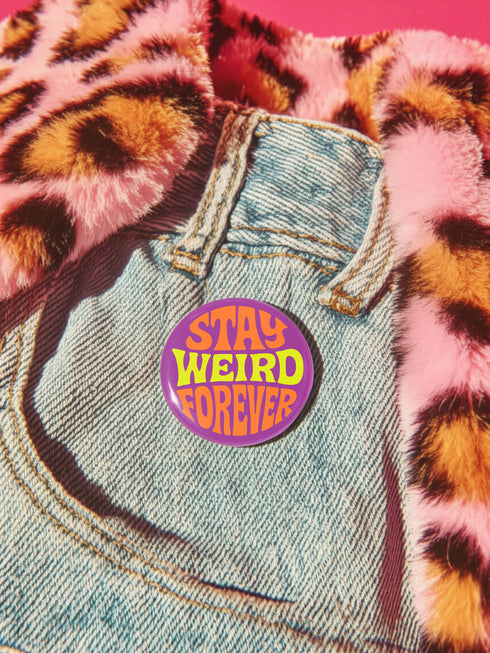 A round metal pin button with a glossy finish and a vibrant purple background. The phrase 'STAY WEIRD FOREVER' is written across the center in a wavy, psychedelic 70s-style font. The words 'STAY' and 'FOREVER' are a bold, bright orange, while the word 'WEIRD' is rendered in a contrasting, electric chartreuse lime green. The button has a smooth, scratch-resistant mylar surface covering the design. The pin is shown in its 2-inch and 3-inch variations, highlighting its circular shape and durable construction.