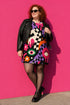 A short-sleeve, above-the-knee t-shirt dress with a relaxed, straight fit and a crew neckline. The dress features a vibrant, chaotic, all-over graphic print. The design includes sections of distorted, wavy black and cream checkerboard, mixed with bold, abstract shapes in bright purple, hot pink, and vivid orange. Scattered throughout the pattern are motifs of hot pink dripping slime, a black bat silhouette, a stylized green eye with a pink iris, and retro pink and purple daisy flowers.