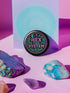 Glossy black pinback button with pastel embroidery-style design that says “Hex the System,” surrounded by vines, stars, and a crescent moon. Displayed against witchy pink and blue backdrops, street pole flyers, and protest props.