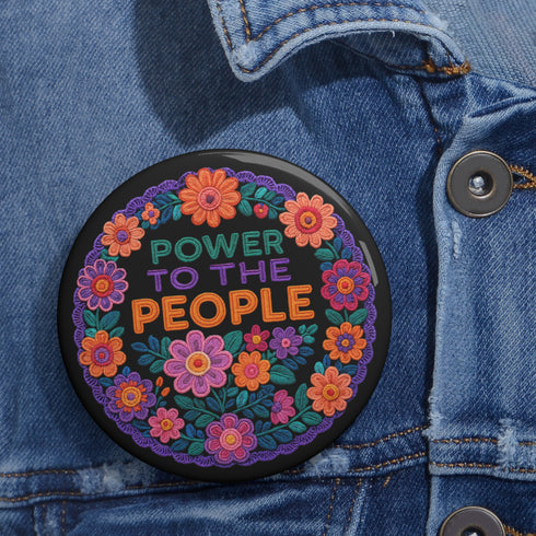 Embroidered-style pinback button featuring the phrase "POWER TO THE PEOPLE" in bold, multicolored satin-stitch letters—green, purple, and orange—framed by a vivid circular floral wreath. Pink, red, and purple flowers with leafy greens radiate around the text. Design has a handcrafted, folk-art feel with a transparent background.