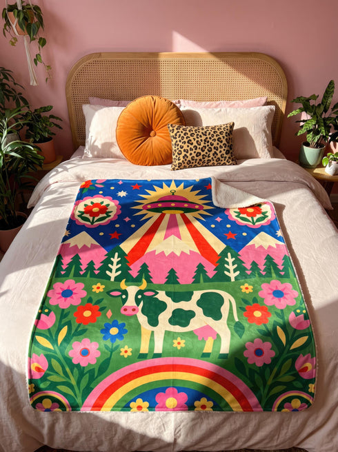 A rectangular velveteen minky blanket with a bright, maximalist design. The top half features a vibrant blue sky with a pink, green, and yellow UFO beaming rays down over hot pink snow-capped mountains. The bottom half shows a grassy green field with a white cow covered in dark spots, surrounded by large, colorful red, pink, and blue retro flowers, pine trees, and a rainbow arch at the very bottom. The reverse side is a fluffy off-white sherpa.