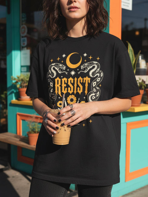 A unisex short sleeve graphic t shirt in dark charcoal black featuring a highly detailed symmetrical medallion print on the chest. In the center, the word RESIST is printed in a bold yellow gothic blackletter font. Two coiled white snakes with fine black scale details frame the text on the left and right. Above the text, a yellow crescent moon is flanked by white and yellow starbursts. Beneath, three yellow marigold flowers sit atop white stems and leafy foliage.