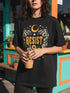 A unisex short sleeve graphic t shirt in dark charcoal black featuring a highly detailed symmetrical medallion print on the chest. In the center, the word RESIST is printed in a bold yellow gothic blackletter font. Two coiled white snakes with fine black scale details frame the text on the left and right. Above the text, a yellow crescent moon is flanked by white and yellow starbursts. Beneath, three yellow marigold flowers sit atop white stems and leafy foliage.