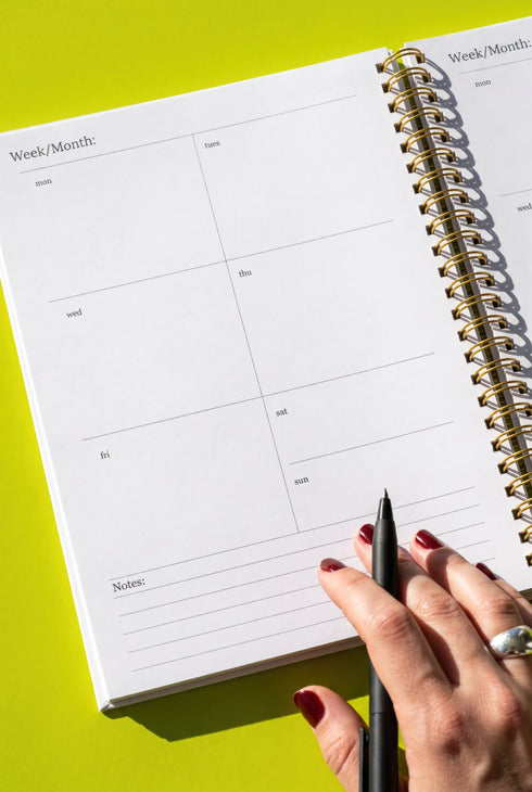 a person's hand holding a black pen, with a spiral-bound notebook open in front of them. The notebook appears to be a daily planner or journal, with blank pages and a calendar section visible. The background is a bright yellow color.