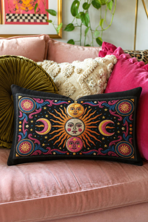 Rectangular 20x12 inch lumbar pillow cover in black featuring printed faux crewel embroidery of a large orange sunburst with a cream face, flanked by pink and yellow crescent moons, a golden moon face above and a pink moon face below, gold stars, blue dots, magenta and blue baroque scroll flourishes, and four corner medallions with golden flowers. Whimsigoth celestial design printed on both sides with a hidden zipper, crafted from soft linen look polyester.