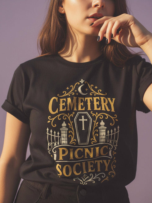 A black unisex crewneck graphic t-shirt featuring an ornate Victorian-inspired emblem printed on the chest in vintage gold and off-white. The centralized, symmetrical design showcases a classic coffin with a cross on the lid, flanked by wrought-iron cemetery gates. Beautiful scrollwork and decorative filigree flourishes surround the bold, retro serif text reading CEMETERY PICNIC SOCIETY. A crescent moon and stars sit at the top, completing this gothic memento mori graphic shirt.
