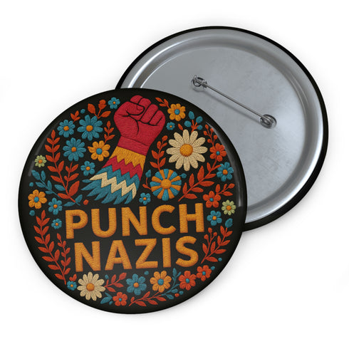 Round black pinback button with bold embroidered-style design featuring a raised red fist, zigzag sleeve, and colorful folk art flowers in orange, teal, yellow, and white. Center text reads “PUNCH NAZIS” in bright golden thread-style lettering. The design is bold, anti-fascist, and maximalist, shown on a glossy pin surface against a pink and purple gradient background. Statement accessory for protest fashion, punk style, and political activism.