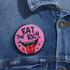 Round pink pinback button featuring a cartoon-style open mouth with sharp teeth, a red tongue, and dripping red details. The bold black text reads “EAT THE RICH” in a hand-drawn style. Glossy black rim with a vibrant, rebellious graphic design.