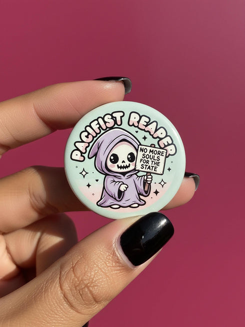Round pinback button featuring a pastel mint green background with a cartoon grim reaper character in a lavender hood holding a protest sign. The design includes bold black outlined lettering reading “Pacifist Reaper” above the figure and small sparkle accents surrounding it. The finish is glossy and smooth, with a printed illustration style on a metal pin badge surface.