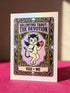 A vertical folded greeting card featuring a pastel goth tarot design titled The Devotion. The central illustration shows a pale devil imp with black wings and horns, holding a red heart in the left hand and a black dagger in the right. The imp has one star eye and one heart eye. The background is lavender purple. A black border frames the scene with teal vines, skulls, gold crescent moons, and roses. The bottom banner reads YOU + ME. The finish is shimmering pearlescent.