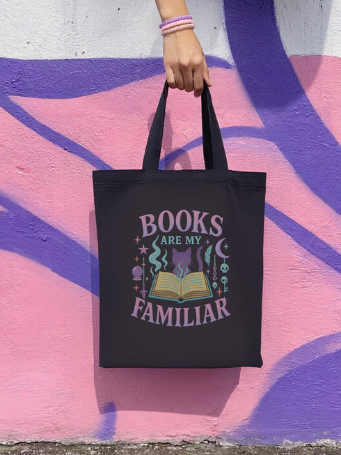 Black cotton tote bag with pastel goth design featuring “Books Are My Familiar” in soft pink, lavender, and mint tones. Design includes an open book with smoke, a silhouetted cat, moons, keys, stars, and magical icons. Shown in front of colorful mural walls and worn by models in alternative fashion. Double-sided print, perfect for witchy readers, soft goths, and literary magic lovers.