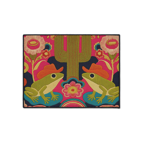 A rectangular polyester doormat with a black trim and non-slip rubber backing. The vibrant, colorful design features a symmetrical illustration on a dark navy blue background. In the center, two green frogs wearing orange cowboy hats sit facing each other. Between them is a tall, olive-green saguaro cactus. The scene is filled with large, psychedelic-style flowers in hot pink, orange, and yellow. The overall aesthetic is funky, retro 70s, maximalist, and trippy, mimicking the look of embroidery.
