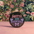Glossy black pinback button featuring pastel-style text that reads “Pretty and Possessed” with a bow, candles, hearts, and sparkles.