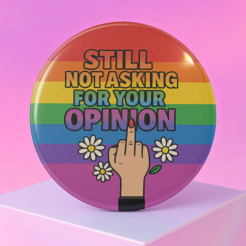 Round glossy pinback button measuring 2.25 inches with a bold rainbow pride flag background. Centered text reads “Still Not Asking For Your Opinion” in black, yellow, blue, and purple letters. Below the text is an illustrated hand with painted red nails giving the middle finger, surrounded by white daisies. The design is printed on tinplate with a scratch- and UV-resistant mylar coating. Button features a strong safety pin backing, perfect for bags, jackets, or pride outfits.