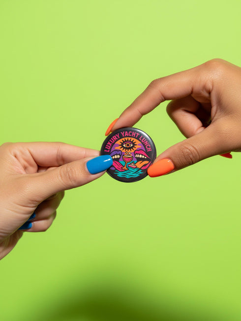 A round 1.75 inch glossy pinback button featuring satirical psychedelic artwork on a black background. The text LUXURY YACHT LUNCH arches over the top in bold neon pink letters. The central design depicts a surreal scene with a stylized third-eye sun, large open mouths with tongues sticking out, a smiling cocktail glass character, and a small white yacht floating on wavy teal water. The color palette includes retro 70s neon pink, orange, purple, and teal. The button has a safety pin backing.