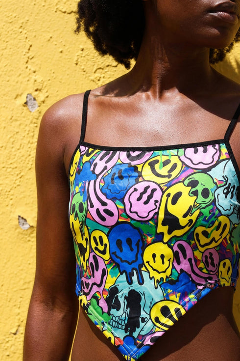A sleeveless women's camisole top featuring a pointed handkerchief hemline often called a bandana top style. The fabric displays a vibrant all-over psychedelic print of distorting and melting smiley faces mixed with melting skulls. The color palette includes bright neon yellow, electric blue, hot pink, and lime green against a multi-colored background. The top features thin black adjustable spaghetti straps and black trim along the straight neckline. The fit is cropped and form-fitting.