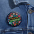 Circular black pinback button with "FUCK THE PATRIARCHY" in orange and mint green text, surrounded by embroidery-style flowers in pink, blue, and red with green foliage and blue border. Folk art feminist protest pin with glossy finish and safety pin backing in 2" and 3" sizes.