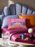 A bright magenta pink rectangular lumbar pillow cover featuring a flat 2D printed faux embroidery design. The printed illusion shows light pink bubble text reading NO THOUGHTS JUST BLANKETS. Below the text is a cartoon pink brain with a happy face, tucked into a blue bed with blue pillows. The brain is covered by a textured-looking orange blanket decorated with light purple daisies. The entire design is a smooth printed trompe l'oeil stitch effect, not real woven or stitched texture.
