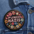 Round pinback metal button featuring an embroidered-style design on a black background. The center showcases bold, stitched-textured lettering in red, blue, and white that reads "PUNCH NAZIS." Surrounding the text is an intricate, folk-inspired floral pattern with multicolored flowers in shades of pink, orange, blue, and white, arranged symmetrically. A decorative zigzag border frames the edge of the button. The backside is silver metal with a secure pin closure.