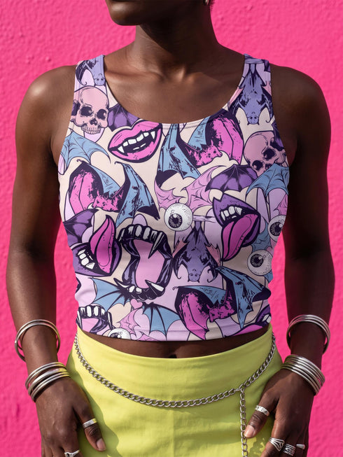 A sleeveless cropped tank top made from stretchy performance knit fabric. The all-over print features a dense, scattered pattern of pastel goth and creepy cute motifs over a light background. Graphics include pink and blue bats in flight, floating eyeballs with blue irises, pink skulls, and glossy vampire lips with sharp white fangs and tongues sticking out. The neckline is a high scoop and the armholes are curved. The fabric comes in either a matte finish or a sparkly silver glitter foil.