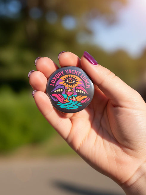 A round 1.75 inch glossy pinback button featuring satirical psychedelic artwork on a black background. The text LUXURY YACHT LUNCH arches over the top in bold neon pink letters. The central design depicts a surreal scene with a stylized third-eye sun, large open mouths with tongues sticking out, a smiling cocktail glass character, and a small white yacht floating on wavy teal water. The color palette includes retro 70s neon pink, orange, purple, and teal. The button has a safety pin backing.